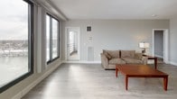 Located at 170 Washington in Haverhill, MA, this single-floor residence offers a thoughtfully designed layout with modern finishes throughout. The kitchen is outfitted with stainless steel appliances—including a refrigerator, electric range, and over-the-range microwave—paired with crisp white shaker cabinetry and light hardwood-style flooring that extends throughout the main living areas. An open peninsula connects the kitchen to a dedicated dining area, which comfortably accommodates a full dining table, making the space ideal for both everyday meals and entertaining. The living room is bright and airy, featuring large floor-to-ceiling windows that invite natural light and offer views of the surrounding cityscape, with access to a balcony. The bedroom is a calm retreat with soft carpeting, ample room for full-sized furniture, and notable architectural beam detailing on the ceiling. A spacious walk-in closet with wire shelving provides generous storage. The bathroom presents a clean, contemporary aesthetic with a white vanity, modern fixtures, and light-toned flooring that complements the overall palette. Neutral gray walls and consistent flooring choices throughout lend the space a cohesive, modern character.