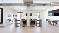 This contemporary single-floor commercial space offers a well-designed open-plan layout that balances functionality with modern style. The interior is defined by crisp white walls, light wood-look tile flooring, and prominent structural columns that divide the space into distinct functional zones while maintaining an open, airy feel.

The main area features dual high-top communal work tables paired with stylish industrial bar stools, ideal for collaborative work or casual meetings. Exposed metallic HVAC ductwork runs along the ceiling, lending an industrial-chic aesthetic that complements the clean, minimalist design. Multiple wall-mounted flat-screen displays are positioned throughout the space.

A comfortable lounge area is anchored by a tufted leather Chesterfield sofa and coordinating armchairs arranged around a low-profile rug, providing a relaxed setting for conversation or informal gatherings. A dedicated refreshment bar counter with dark countertops and bar seating accommodates a coffee station, making it a convenient amenity for occupants.

The entry area features a generously sized glass door and expansive floor-to-ceiling windows that flood the interior with natural light and provide views of the surrounding landscaping. A small seating nook near the entrance, furnished with a pair of modern accent chairs and a round side table, creates a welcoming reception area. Decorative wall art is thoughtfully placed throughout, adding personality to the professional environment.