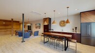 Located on Village Green Boulevard in Aiken, South Carolina, this single-floor community amenity space offers a generously sized, thoughtfully designed environment for residents to gather, dine, work out, and connect. Light wood-look tile flooring flows throughout, unifying the open-concept layout and enhancing the bright, welcoming atmosphere created by recessed lighting and large entry doors that invite natural light deep into the space.

The expansive main lounge serves as the heart of the facility, featuring sleek black steel columns, comfortable seating areas with plush blue chairs and leather sofas, and a built-in media center with shelving. A striking wooden staircase with black metal railings adds architectural interest, while a sliding barn door set against a rich wood-paneled wall adds warmth and character.

The dining area is a standout feature, anchored by a solid wood bar-height communal table with industrial-style seating, illuminated by a trio of warm gold pendant lights and accented by a decorative sunburst wall piece. Dark wood cabinetry and a wet bar complete the sophisticated setting.

The exercise room is well-appointed with cardio equipment, a ceiling fan, and vibrant landscape murals that energize the space. A separate, softly painted dining or meeting room provides a quieter setting with a conference table and decorative wall accent, ideal for small gatherings or private use.