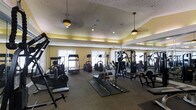 This fitness center is a thoughtfully designed, single-floor facility built to accommodate a full range of exercise needs. The space features warm yellow walls accented by a tray ceiling with recessed lighting and decorative pendant light fixtures, creating a bright and inviting atmosphere. A textured ceiling adds a classic touch to the overall aesthetic.

The gym is generously outfitted with a diverse selection of Cybex-brand strength training machines, free weight benches, and a variety of cardio equipment including treadmills, ellipticals, and stationary bikes. Rubber floor mats are positioned beneath the weight equipment to provide safety and stability during use. Large mirrors line one wall, enhancing the sense of space and allowing users to monitor their form during workouts.

Natural light filters in through multiple windows fitted with blinds, supplementing the overhead lighting and contributing to an open, energizing environment. The layout is organized to allow comfortable movement between stations, making it practical for simultaneous use by multiple individuals. Overall, the space delivers a clean, functional, and well-maintained environment suited for a wide variety of fitness routines.