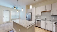 Located in Friendswood, Texas, this single-floor residence offers a thoughtfully designed layout with contemporary finishes throughout. The kitchen serves as the heart of the home, showcasing a large granite-topped island with pendant lighting overhead, light wood-tone cabinetry extending to the ceiling, a subway tile backsplash, and a full suite of stainless steel appliances including a French door refrigerator, electric range, microwave, and dishwasher. Wood-look flooring runs through the main living and kitchen areas, adding warmth and continuity to the open layout. Ceiling fans provide comfort throughout the common areas, which also benefit from natural light through slatted blinds. The bedrooms feature plush neutral carpeting and windows that invite natural light, creating a calm and restful atmosphere. One versatile room includes a built-in granite-topped desk with cabinetry and overhead shelving — ideal as a home office or study. A hallway connecting the private spaces leads to a well-appointed bathroom featuring a glass-enclosed walk-in shower with tiled walls. Clean white trim, crown molding details, and a cohesive neutral palette tie the entire home together, resulting in a modern yet comfortable living environment.
