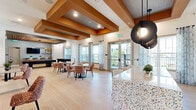Located at 904 S Lake St in Leesburg, Florida, this thoughtfully designed single-floor community clubhouse offers a welcoming and versatile gathering space with a refined, contemporary aesthetic. The open layout seamlessly connects multiple activity zones, creating a fluid social environment bathed in natural light from large windows dressed with patterned drapery.

The fully equipped kitchen serves as a visual centerpiece, showcasing gray shaker cabinetry, open shelving, a vertical tile backsplash, stainless steel appliances, and a generous island topped with striking terrazzo-style countertops. Three oversized globe pendant lights in a matte black finish hang above the island, which is lined with modern bar-height seating — ideal for casual gatherings or catered events.

Adjacent to the kitchen, a spacious game room features a full-size billiards table set beneath a dramatic circular chandelier and slatted wood ceiling detail. Light wood-toned flooring flows throughout, complemented by warm orange accent beams that add architectural character across the ceiling.

A dedicated lounge and dining area offers padded banquette seating along a teal accent wall adorned with framed local artwork, alongside patterned upholstered chairs arranged around communal tables. The space combines comfort and style, making it well-suited for both relaxed socializing and organized community events.