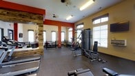 This fitness center offers a well-appointed, single-floor workout space designed to support a full range of exercise routines. The interior is characterized by warm yellow and bold terracotta-red accent walls, creating an energizing environment that motivates activity. A distinctive rustic wood accent column adds a touch of unique architectural character to the space.

The flooring throughout is durable rubber, ideal for high-impact workouts and the placement of heavy equipment. The center is outfitted with a comprehensive selection of strength-training machines, including a multi-station cable and weight machine, as well as dedicated benches for targeted exercises. A designated cardio area is visible toward the rear, housing additional equipment such as stationary bikes and stability balls.

Natural light floods the space through multiple large windows fitted with white horizontal blinds, contributing to an open and airy feel. Ceiling fans provide comfortable air circulation, while fluorescent lighting ensures the space remains well-lit at all times. A railed partition separates functional zones within the facility, offering an organized layout that accommodates multiple users simultaneously. Overall, the fitness center presents a clean, practical, and welcoming environment suited for a broad range of fitness goals.