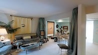 This two-floor space presents a distinctly eclectic and artistic interior aesthetic. The main living area is furnished with vintage-style seating, including a tufted sofa and ornate accent chairs arranged around a distressed blue coffee table, creating a curated, bohemian atmosphere. Muted sage-green floor-length curtains frame the room, adding softness and color contrast against the neutral walls and light carpeted flooring. Artwork and framed pieces are displayed throughout, contributing to the space's gallery-like character, with wall hangings and decorative objects lending a deeply personal, creative sensibility. An open archway connects the living area to an adjacent dining space, which features a light-painted table and chairs alongside additional wall-mounted mirrors and artwork. The dining area is painted in a soft aqua-blue tone, providing a subtle color shift that visually distinguishes the two zones while maintaining a cohesive overall palette. Natural and recessed lighting illuminate both areas, keeping the interior bright. The layered mix of vintage furniture, curated art pieces, and rich textiles throughout gives the space a warm, lived-in, and highly individualistic character.