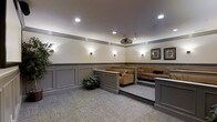 This well-appointed screening room is designed for comfortable, private viewing experiences. The space features a tiered layout, with a raised platform at the rear accommodating plush, tan upholstered sofas and seating arrangements that ensure clear sightlines from every seat. The lower level provides additional seating flexibility, separated by a low partition wall finished in grey.

The room's décor balances elegance with functionality. Grey wainscoting lines the lower portion of the walls, complemented by warm cream-toned upper walls adorned with framed artwork. Recessed ceiling lighting and wall-mounted sconces provide warm, ambient illumination throughout the room, creating a cozy atmosphere suited to screenings. A ceiling-mounted projector is centrally positioned, ready to deliver a full cinematic experience.

The flooring throughout is a neutral, patterned carpet that adds comfort underfoot while contributing to the room's acoustic quality. Crown moulding runs along the ceiling perimeter, adding a refined architectural touch. Decorative indoor plants are placed around the room, softening the overall aesthetic and adding a welcoming, lived-in feel to this purpose-built entertainment space.