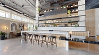 Located at 2990 Longwater Blvd in Kissimmee, Florida, The Maxwell is a striking communal space that blends industrial architecture with refined, contemporary design. The single-floor layout spans a generous footprint, anchored by dramatic double-height ceilings with exposed concrete columns, open ductwork, and expansive floor-to-ceiling windows that flood the interior with abundant natural light and offer sweeping outdoor views.

At the heart of the space is a stunning long bar counter topped with white marble, complemented by warm light wood cabinetry and sleek dark bar stools — an ideal setting for socializing or working. A dedicated lounge area features rich brown leather sectional sofas, sculptural white boucle chairs, and layered geometric rugs, all bathed in sunlight streaming through the oversized window walls.

A dramatic living green wall serves as a striking focal point, climbing toward the ceiling alongside warm slatted wood paneling and lush potted plants that reinforce a connection to nature throughout. Display shelving stocked with curated décor and greenery lines the upper walls, adding visual depth and warmth.

The polished concrete floors, pendant lighting, and carefully selected furnishings contribute to an atmosphere that is simultaneously modern and inviting — a sophisticated, amenity-rich space designed for comfort and community.