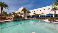 Located at 2500 Edwards Dr in Fort Myers, Florida, this expansive outdoor pool complex offers a resort-style experience in the heart of Southwest Florida. The centerpiece of the space is a generously sized swimming pool with crystal-clear turquoise water, featuring designated lap lanes for fitness-oriented guests alongside open recreational areas. The pool is flanked by decorative tiled accent walls with a blue-and-white mosaic pattern, adding a distinctive architectural touch to the environment.

Surrounding the pool, a broad sun deck is furnished with lounge chairs, tables, and vibrant blue market umbrellas, providing ample shaded and open-air relaxation areas. Tall, mature palm trees are thoughtfully interspersed throughout the deck, enhancing the tropical ambiance and offering natural shade under the bright Florida sun. Lush tropical plantings and manicured hedges define the perimeter of the space, creating a sense of privacy and seclusion.

The complex is set against the backdrop of a modern, multi-story white building with large floor-to-ceiling windows and a high-rise tower, indicating a substantial hospitality or commercial facility. A secondary staircase and elevated deck area are also visible, suggesting additional amenity spaces. The overall atmosphere is bright, open, and inviting, well-suited to the warm Fort Myers climate.