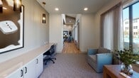 Located at 19770 Clay Rd in Katy, Texas, this single-floor community amenity space offers a thoughtfully designed environment that blends modern aesthetics with functional layouts. The space features a welcoming open lounge area with soaring vaulted ceilings, warm natural wood-look flooring, and an abundance of natural light streaming through floor-to-ceiling windows dressed in sheer neutral drapery. The lounge is furnished with comfortable accent chairs and anchored by a wall-mounted display, creating an inviting gathering area with an airy, open ambiance.

A dedicated business center provides a productive workspace equipped with a continuous built-in white countertop desk, ample storage cabinetry, ergonomic seating, and stylish pendant lighting with dark cylindrical shades. Framed wall art adds a polished touch, while direct exterior access makes the area easily accessible.

Transitional spaces connect the zones seamlessly, with plush carpet in the lounge and work areas contrasting the gleaming hardwood-style flooring of the main corridor. Cozy windowed nooks outfitted with cushioned armchairs and side tables offer quiet retreats bathed in outdoor views of the surrounding community. Recessed lighting throughout ensures consistent, well-distributed illumination, completing the clean and contemporary feel of this versatile communal space.