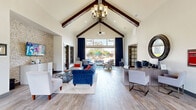 Located along Highway 105 W in Montgomery, Texas, this single-floor community space offers a thoughtfully designed layout encompassing a living room, kitchen, and office. The entrance foyer greets visitors with soaring vaulted ceilings accented by exposed dark wood beams, light wood-look tile flooring, and a grand wooden front door flanked by glass sidelights that invite abundant natural light. The expansive living room continues the vaulted ceiling aesthetic, featuring dramatic floor-to-ceiling windows dressed in deep navy curtains that frame views of the outdoor courtyard and pool area. A ceiling fan anchors the space above, while light hardwood-style flooring extends throughout. A secondary lounge area provides additional seating with recessed lighting, built-in display shelving, and large windows overlooking the exterior. The kitchen is outfitted with rich, dark espresso cabinetry, granite countertops, stainless steel appliances including a full-size refrigerator, a central island with bar seating, and stylish pendant lighting overhead — all complemented by wood-look tile flooring. The dedicated office is accessed through elegant glass-paned French doors and features built-in desk workstations, carpeted flooring, crown molding, and natural light from a window with decorative drapery, accommodating multiple workstations for resident use.