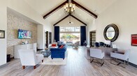 Located along Highway 105 W in Montgomery, Texas, this single-floor community space offers a thoughtfully designed layout encompassing a living room, kitchen, and office. The entrance foyer greets visitors with soaring vaulted ceilings accented by exposed dark wood beams, light wood-look tile flooring, and a grand wooden front door flanked by glass sidelights that invite abundant natural light. The expansive living room continues the vaulted ceiling aesthetic, featuring dramatic floor-to-ceiling windows dressed in deep navy curtains that frame views of the outdoor courtyard and pool area. A ceiling fan anchors the space above, while light hardwood-style flooring extends throughout. A secondary lounge area provides additional seating with recessed lighting, built-in display shelving, and large windows overlooking the exterior. The kitchen is outfitted with rich, dark espresso cabinetry, granite countertops, stainless steel appliances including a full-size refrigerator, a central island with bar seating, and stylish pendant lighting overhead — all complemented by wood-look tile flooring. The dedicated office is accessed through elegant glass-paned French doors and features built-in desk workstations, carpeted flooring, crown molding, and natural light from a window with decorative drapery, accommodating multiple workstations for resident use.