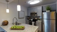 This single-floor residence showcases a thoughtfully designed interior with a strong emphasis on modern aesthetics and functional living. The kitchen serves as a standout feature, outfitted with sleek grey shaker-style cabinetry that provides ample storage while contributing to the space's contemporary feel. Stainless steel appliances, including a refrigerator, electric range, and over-the-range microwave, offer both style and practicality. The granite countertops, featuring a light, speckled finish, extend generously across the workspace and into a breakfast bar or island area, ideal for casual dining or meal prep. A grey subway tile backsplash adds texture and visual interest behind the range, tying together the cool-toned color palette throughout the kitchen. Overhead, a large flush-mount ceiling light provides bright, even illumination, complemented by a modern pendant light that adds a warm, focused glow. The neutral grey walls create a clean, versatile backdrop, while decorative touches lend a welcoming, homey ambiance to the space. Overall, the kitchen exudes a polished, move-in-ready character with a cohesive design that balances comfort and modern style.