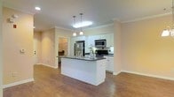 This single-floor residence showcases an open-concept layout that seamlessly connects the kitchen and living areas. The kitchen is a standout feature, equipped with crisp white cabinetry extending to the ceiling, stainless steel appliances including a refrigerator, microwave, and range, and a dark tile backsplash that provides a striking contrast. A central island with a dark countertop and an integrated sink serves as both a functional workspace and a casual gathering point, illuminated by elegant pendant lighting overhead. Crown molding runs throughout the space, adding a refined architectural detail that elevates the overall aesthetic. The warm hardwood-style flooring flows continuously across the main living area, contributing to a cohesive and inviting atmosphere. Neutral-toned walls enhance the brightness of the space, complemented by recessed and pendant lighting that ensures even illumination throughout. A doorway visible from the kitchen leads to an additional room, suggesting a well-connected and practical floor plan. The overall ambiance is clean, modern, and well-appointed, combining functional design with tasteful finishes.
