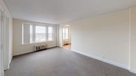This single-floor space showcases a thoughtful layout with a blend of classic architectural character and functional design. The floor plan, visible in the 3D model, reveals a series of interconnected rooms arranged in an L-shaped configuration, providing a natural flow between living areas.

The main living area is generously proportioned, featuring neutral wall-to-wall carpeting, bright cream-colored walls, and a striking arched doorway that leads into an adjoining room. Multiple windows allow ample natural light to fill the space, complemented by cast-iron radiators that underscore the building's classic heritage.

Through the arch lies a room finished with rich hardwood floors in a warm honey tone, adding character and warmth. This room features decorative chair-rail molding along the walls, a pendant light fixture with a frosted glass shade suspended from the ceiling, and a radiator positioned beneath the windows — details that speak to the space's period charm.

Additional carpeted rooms branch off from the main areas, each finished with the same cream palette and white baseboard trim, offering clean, versatile spaces. Doors throughout feature traditional paneled designs consistent with the building's architectural style. Crown molding runs along the ceilings, tying the rooms together with a refined, cohesive aesthetic throughout the entire unit.