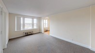 This single-floor space showcases a thoughtful layout with a blend of classic architectural character and functional design. The floor plan, visible in the 3D model, reveals a series of interconnected rooms arranged in an L-shaped configuration, providing a natural flow between living areas.

The main living area is generously proportioned, featuring neutral wall-to-wall carpeting, bright cream-colored walls, and a striking arched doorway that leads into an adjoining room. Multiple windows allow ample natural light to fill the space, complemented by cast-iron radiators that underscore the building's classic heritage.

Through the arch lies a room finished with rich hardwood floors in a warm honey tone, adding character and warmth. This room features decorative chair-rail molding along the walls, a pendant light fixture with a frosted glass shade suspended from the ceiling, and a radiator positioned beneath the windows — details that speak to the space's period charm.

Additional carpeted rooms branch off from the main areas, each finished with the same cream palette and white baseboard trim, offering clean, versatile spaces. Doors throughout feature traditional paneled designs consistent with the building's architectural style. Crown molding runs along the ceilings, tying the rooms together with a refined, cohesive aesthetic throughout the entire unit.