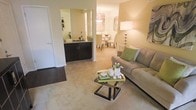 This single-floor residence offers a thoughtfully designed open-concept layout that seamlessly connects the living, dining, and kitchen areas into one cohesive living space. The living room is anchored by plush carpeting, a contemporary glass-and-metal coffee table, and a comfortable sofa accented with decorative throw pillows, creating a cozy yet stylish atmosphere. Warm, neutral tones on the walls complement the soft lighting provided by an elegant floor lamp, adding to the inviting ambiance. Adjacent to the living area, the kitchen features dark cabinetry with sleek hardware and stone countertops, offering a striking contrast to the lighter surroundings. A pass-through counter connects the kitchen to the main living space, enhancing the open flow. The dining area, visible in the background, is bathed in warm light from a pendant fixture above, and is furnished with a glass-top dining table and coordinating chairs — ideal for both casual meals and entertaining. Hardwood-style flooring in the kitchen transitions gracefully to soft carpet in the living areas, adding texture and visual interest. The overall design strikes a balance between functionality and comfort, making this a welcoming and practical space.