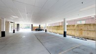 This covered parking facility presents a well-maintained, single-level structure designed for efficient vehicle accommodation. The space features a clean white drop-panel ceiling that runs the full expanse of the facility, providing a bright and open atmosphere. Structural columns are strategically positioned throughout, with protective bollards installed at their bases to safeguard both vehicles and the building's integrity. The smooth concrete floor is clearly marked with parking space delineations, ensuring organized and orderly use of the available area. Along one side of the facility, a wooden privacy fence defines the perimeter, adding a degree of enclosure while complementing the surrounding brick architecture visible beyond. The open sides of the structure allow for ample natural light to penetrate the interior, creating a welcoming and well-lit environment without the need for extensive artificial lighting during daylight hours. The overall design balances functionality and practicality, offering a spacious and accessible parking solution. The facility's clean lines, neutral color palette, and thoughtful layout make it a highly functional space suitable for a variety of residential or commercial parking needs.