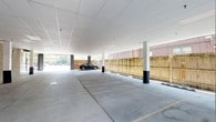 This covered parking facility presents a well-maintained, single-level structure designed for efficient vehicle accommodation. The space features a clean white drop-panel ceiling that runs the full expanse of the facility, providing a bright and open atmosphere. Structural columns are strategically positioned throughout, with protective bollards installed at their bases to safeguard both vehicles and the building's integrity. The smooth concrete floor is clearly marked with parking space delineations, ensuring organized and orderly use of the available area. Along one side of the facility, a wooden privacy fence defines the perimeter, adding a degree of enclosure while complementing the surrounding brick architecture visible beyond. The open sides of the structure allow for ample natural light to penetrate the interior, creating a welcoming and well-lit environment without the need for extensive artificial lighting during daylight hours. The overall design balances functionality and practicality, offering a spacious and accessible parking solution. The facility's clean lines, neutral color palette, and thoughtful layout make it a highly functional space suitable for a variety of residential or commercial parking needs.