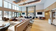 Located at 5505 W Northgate Rd in Rogers, Arkansas, this generously sized single-floor amenity space offers a sophisticated and welcoming environment designed for both relaxation and socializing. The open-plan layout is anchored by soaring double-height ceilings in the main lounge area, where a striking sculptural gold chandelier serves as a dramatic focal point above plush, neutral-toned sectional seating arranged on a large patterned area rug.

Light oak hardwood flooring runs throughout, unifying the various zones seamlessly. A built-in wet bar with marble countertops, pendant lighting, and open cabinetry provides an elegant focal point at one end of the room, complemented by bar seating and a wall-mounted flat-screen television. Warm wood paneling lines portions of the walls, adding texture and refinement to the interior palette.

A more intimate lounge area features coffered ceilings with rich dark wood beams, textured blue-gray wall treatments, curated artwork, and comfortable seating — creating a quieter retreat within the larger space. Additional zones accommodate a billiards table and casual seating arrangements. Floor-to-ceiling black-framed glass doors and windows flood the space with natural light and offer direct views and access to an outdoor pool and courtyard beyond.