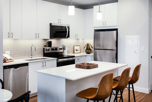 Cocina de la colección Design I con electrodomésticos de acero inoxidable, gabinetes blancos, protector contra salpicaduras de azulejos grises, iluminación debajo de los gabinetes, encimeras de cuarzo blanco y pisos de superficie dura. - Avalon Quincy Adams