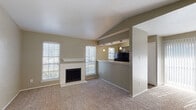 Located at 5555 Spring Valley Rd in Dallas, Texas, this single-floor residence offers a well-organized layout with distinct living, kitchen, bedroom, and bathroom spaces. The living room is a standout area, featuring vaulted ceilings with angled beams that lend an airy, open feel. A brick-surround fireplace with a white mantel serves as the room's focal point, flanked by multiple windows dressed with white blinds that allow generous natural light to fill the carpeted space. An open pass-through connects the living area to the kitchen, which is equipped with black appliances including a refrigerator and range, light-toned countertops, a stainless steel sink, and wood-look flooring. A window above the kitchen area provides additional natural light. The bedroom areas are carpeted and neutral in tone, with one room featuring a sliding glass door draped in sheer curtains that invite soft, diffused light. Additional bedrooms include windows with blinds, maintaining a bright and comfortable atmosphere throughout. The bathroom is fitted with a combined tub and shower with tiled surround, brushed nickel hardware and towel bar, and wood-look flooring. Neutral warm-beige wall tones are consistent throughout the residence, contributing to a cohesive and calm overall ambiance.