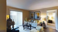 This single-floor residence offers a thoughtfully designed living area that combines comfort and contemporary style. The living room serves as the focal point of the space, showcasing warm, earthy tones on the walls that create an inviting and cozy atmosphere. A large sliding glass door fitted with vertical blinds allows an abundance of natural light to fill the room while providing access to an outdoor area.

The living room is centered around a plush, light gray sofa accented with decorative throw pillows in mustard yellow and neutral tones, complemented by a coordinating ottoman. A dark-finished, round coffee table anchors the seating area, which is further defined by a patterned area rug beneath. A side table with a warm-toned lamp adds ambient lighting to the space.

Wall décor includes two large framed artworks featuring abstract compositions in gold, gray, and blue, adding a curated, gallery-like touch to the room. The smooth white ceiling and clean crown molding contribute to the polished, well-maintained aesthetic of the space. Overall, the living area presents a balanced blend of warmth, natural light, and modern design sensibility.
