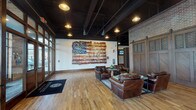 This single-floor community space presents a compelling mix of design styles across several distinct areas. The main lounge entrance makes a bold statement with its exposed dark ceiling beams, pendant lighting, and warm hardwood floors. A large-scale mixed-media American flag artwork anchors one wall, while a seating arrangement of rich leather club chairs and a coffee table creates an inviting social hub. Large glass-paned entry doors allow natural light to flow through the space.

An adjacent sitting area transitions to a more rustic aesthetic, centered around a floor-to-ceiling stacked stone fireplace with a substantial wood mantel. French doors in this area open to an outdoor space, providing additional natural light and exterior access. Neutral upholstered seating complements the organic textures of the stonework.

A third gathering area offers a bright, transitional design featuring high ceilings, elegant drum-shade chandeliers, and a soft neutral color palette. Cream-toned sofas and wingback chairs are arranged alongside a dining table, creating a versatile space for both casual lounging and small gatherings. Black-framed French doors line the walls, connecting this area to adjacent spaces and maintaining a sense of openness throughout.