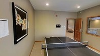 This single-floor space is designed for recreational use and casual enjoyment. The main area is anchored by a full-size ping pong table, making it well-suited for active leisure and social gatherings. The walls are finished in a neutral taupe tone, contributing to a relaxed and welcoming atmosphere. Recessed ceiling lighting provides even, functional illumination throughout the room. A large, bold piece of framed artwork adorns one wall, adding a vibrant visual focal point with its colorful, abstract design. A ball holder is conveniently mounted on the wall near the table, keeping equipment organized and accessible. The space features carpeted flooring with warm wood baseboards that complement the overall neutral palette. An open doorway connects this recreational room to an adjacent lounge area, which is visible in the background and features hardwood-style flooring, a cushioned chair, and warm wood-toned doors—suggesting a seamless flow between activity and relaxation zones. The layout is straightforward and functional, with ample open floor space surrounding the ping pong table to allow for comfortable gameplay.