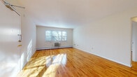 This single-floor residence offers a clean, well-maintained interior defined by warm, polished hardwood floors that extend throughout the living areas and bedrooms, reflecting natural light and lending the space a bright, welcoming character.

The spacious living room is a standout feature, offering generous proportions, crisp white walls, and multiple windows that bathe the room in sunlight. Baseboard heating and a wall-mounted air conditioning unit ensure year-round comfort. A dedicated dining area adjoins the living space, illuminated by a classic chandelier and a window with an outdoor view, making it an ideal spot for meals and gatherings.

The kitchen is equipped with warm honey-toned wood cabinetry, granite-style countertops, a tile backsplash, and white appliances including a gas range and over-the-range microwave. Tile flooring completes the practical and tidy aesthetic of the space.

The bedroom offers a tranquil retreat with the same gleaming hardwood floors and ample wall space. The bathroom is tastefully finished with large-format neutral ceramic tile surrounding a full soaking tub and shower combination, complemented by a white vanity with storage and a mirrored medicine cabinet above.

Throughout the residence, the design emphasizes clean lines, natural light, and functional simplicity.