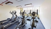 This fitness center is a comprehensively equipped exercise facility designed to support a full range of workout routines. The space is organized into distinct functional zones, making efficient use of the available floor area. The cardio section is prominently positioned along a wall of large windows, which allow generous natural light to fill the room and offer views of the surrounding neighborhood, creating an open and energizing atmosphere during workouts.

The cardio zone houses multiple treadmills, elliptical cross-trainers, and stationary bikes, all arranged in orderly rows atop durable rubber flooring that covers the entire facility. The strength and free-weight area is located toward the rear of the space, defined by a partial partition wall. This section features a dumbbell rack with a mirrored back wall, metal storage cabinetry, and an adjustable weight bench, providing a dedicated environment for resistance training. Exposed ductwork and red-piped sprinkler lines run along the ceiling, contributing an industrial character to the space.

Flush-mounted fluorescent light fixtures provide consistent overhead illumination throughout. Bulletin boards, a sanitizing station, and a wall clock reflect a practical, user-focused design. A wood-framed entry door with a sidelight window completes the layout.
