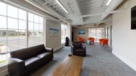 Located at 27 Davis St in Warren, Rhode Island, this single-floor commercial space offers a thoughtfully designed open-plan layout that balances collaborative functionality with a welcoming ambiance. The exposed ceiling structure, finished in white and accented by industrial-style linear lighting and visible mechanical systems, lends an airy, modern-industrial character throughout.

The space is organized into distinct functional zones. A lounge area features dark leather sofas and armchairs arranged around a low wood coffee table, ideal for informal gatherings or casual meetings. Adjacent to this is a second seating zone furnished with vibrant orange upholstered tub chairs paired with round accent tables, adding a bold pop of color against the neutral, warm-toned walls. A dedicated meeting area includes a substantial dark wood conference table surrounded by metal-frame chairs, suitable for more formal discussions. A wall-mounted flat-screen television is positioned to serve both the lounge and meeting areas.

Patterned carpet tiles in muted blue-gray tones cover the floors throughout, providing visual texture while maintaining a professional appearance. Large windows along the perimeter admit generous natural light and provide views of the surrounding Warren streetscape. Coastal-themed artwork adorns the walls, complementing the overall calm, professional atmosphere of the space.
