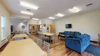 This single-floor community space is designed to accommodate a variety of group activities and social gatherings within an open, welcoming layout. The main common area features warm hardwood-style flooring throughout, bright fluorescent lighting, and neutral-toned walls that create a clean, functional atmosphere. Multiple rectangular dining tables with light wood tops and ladder-back chairs are arranged throughout the space, providing ample seating for groups of various sizes. A dedicated lounge area offers plush blue sofas arranged around a coffee table, with a wall-mounted flat-screen television positioned for easy viewing. A well-stocked bookshelf along one wall adds a casual reading nook to the environment. French doors along one side of the room allow natural light to filter in and provide views of an outdoor green area. A separate vending alcove features a recessed tray ceiling with recessed lighting, large windows, a round dining table, and a Pepsi vending machine. The fully equipped galley-style kitchen includes warm wood cabinetry, a double sink, a full-size refrigerator, a range with an overhead microwave, a dishwasher, and tile flooring, making the space suitable for preparing and serving meals to larger groups.