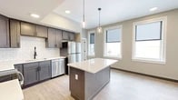 Located at 8 Bank Pl in Utica, NY, this thoughtfully designed 2-bedroom, 1-bathroom unit delivers a modern aesthetic across a single floor. The open-concept kitchen serves as the heart of the home, showcasing dark gray shaker-style cabinetry, crisp white quartz countertops, a classic subway tile backsplash, and a full suite of stainless steel appliances including a range, microwave, dishwasher, and refrigerator. A central kitchen island with pendant lighting above adds both functionality and visual appeal, while light wood-tone flooring ties the space together seamlessly. Large windows flood the kitchen and living area with natural light and offer views of the surrounding Utica streetscape. Both bedrooms are carpeted and painted in a calming sage green, each offering a dedicated closet for ample storage. The bathroom features neutral tile flooring and an eye-catching exposed brick accent wall, blending industrial character with contemporary design. Black hardware accents throughout add a cohesive, modern touch. The layout flows efficiently, making excellent use of the available footprint while maintaining a clean, bright, and welcoming ambiance throughout.