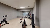 Located at 6325 Victoria Ave in St. Louis, Missouri, this dedicated fitness center offers a well-appointed, single-floor exercise space designed for a comprehensive workout experience. The room features a clean, modern aesthetic with neutral gray walls, light wood-tone flooring, and bright overhead fluorescent lighting that keeps the space well-illuminated throughout.

A defining architectural feature is the large wall-spanning mirror, which visually expands the room and allows users to monitor their form during exercise. The fitness area is equipped with multiple stationary cycling bikes, providing ample space for spin-style workouts. A full dumbbell rack offers a graduated range of free weights, while a medicine ball tower stocked with a colorful variety of weighted balls supports functional training and core work. A Power Systems aerobic step platform rounds out the strength and conditioning equipment.

The space is thoughtfully organized to allow freedom of movement, with equipment arranged along the perimeter and open floor space in the center. A large glass partition wall separates the main workout area from an adjacent corridor, where a water fountain is conveniently accessible. The overall ambiance is clean, functional, and purposeful, making this fitness center a practical and well-equipped facility for a wide range of exercise routines.