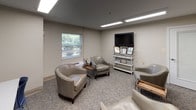 Located at 500 Harry S Truman Dr in Largo, Maryland, this single-floor professional space is thoughtfully designed to accommodate both relaxed interaction and focused work. The main lounge area features plush, silver-toned barrel chairs arranged to encourage conversation, set atop patterned carpet that adds texture and warmth to the neutral color palette. Soft beige walls and recessed linear fluorescent lighting create a clean, well-lit environment throughout. A flat-screen television mounted above a white display shelf with decorative accents provides an inviting focal point in the lounge, while framed artwork depicting nature scenes adds a calming aesthetic to the walls. A window fitted with horizontal blinds allows natural light to filter in and offers views of the surrounding greenery outside. A wall-mounted PTAC unit ensures climate comfort year-round. Adjacent to the lounge, a dedicated workstation area features a long white desk paired with blue task chairs, illuminated by skylight-style ceiling panels. A slate-blue accent wall anchors this zone, further distinguished by framed botanical prints. A frosted-glass interior door connects the areas, maintaining an open yet organized flow. This space is well-suited for professional consultations, client meetings, or collaborative work sessions.