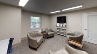 Located at 500 Harry S Truman Dr in Largo, Maryland, this single-floor professional space is thoughtfully designed to accommodate both relaxed interaction and focused work. The main lounge area features plush, silver-toned barrel chairs arranged to encourage conversation, set atop patterned carpet that adds texture and warmth to the neutral color palette. Soft beige walls and recessed linear fluorescent lighting create a clean, well-lit environment throughout. A flat-screen television mounted above a white display shelf with decorative accents provides an inviting focal point in the lounge, while framed artwork depicting nature scenes adds a calming aesthetic to the walls. A window fitted with horizontal blinds allows natural light to filter in and offers views of the surrounding greenery outside. A wall-mounted PTAC unit ensures climate comfort year-round. Adjacent to the lounge, a dedicated workstation area features a long white desk paired with blue task chairs, illuminated by skylight-style ceiling panels. A slate-blue accent wall anchors this zone, further distinguished by framed botanical prints. A frosted-glass interior door connects the areas, maintaining an open yet organized flow. This space is well-suited for professional consultations, client meetings, or collaborative work sessions.