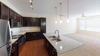 This single-story residence offers a thoughtfully designed layout that combines functionality with modern finishes. The kitchen serves as the centerpiece of the home, showcasing rich dark wood cabinetry that extends to the ceiling, providing ample storage throughout. Granite countertops with a speckled pattern extend across both the perimeter counters and a large island that doubles as a breakfast bar. A double stainless steel sink is set into the island, complemented by a sleek gooseneck faucet. Stainless steel appliances, including a refrigerator, electric range, and built-in microwave, complete the kitchen's polished look. Pendant lighting hangs elegantly above the island, while track lighting illuminates the cooking area. Hardwood flooring anchors the kitchen and flows into the adjacent living area. The bedroom is a comfortable, carpeted space bathed in natural light from a broad window dressed with horizontal blinds. A ceiling fan with an integrated light fixture adds both comfort and charm. The bathroom features a dark-stained vanity with a clean white countertop and single-basin sink, framed by a large mirror and modern multi-bulb vanity lighting above. Textured walls and neutral tones throughout the home create a warm and cohesive ambiance.