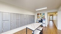 Located at 601 Orange Ave NE in Roanoke, Virginia, this single-floor community amenity space is thoughtfully designed to serve residents with a clean, modern aesthetic. The layout is divided into several functional areas, all unified by warm wood-tone flooring and crisp white walls that create a bright, welcoming atmosphere.

The mail center is a standout feature, occupying a full wall with an extensive array of individual numbered mailboxes in brushed silver, accommodating a large number of residents. A marble-top communal table with stools sits in the center of this area, providing a convenient surface for sorting mail or packages. Adjacent to the mailboxes, a utility station includes a microwave and water cooler, alongside a bulletin board displaying community notices and information.

The lounge area offers a comfortable seating arrangement with a neutral-toned loveseat, a teal accent chair, and a round coffee table set atop a patterned area rug. A wall-mounted flat-screen TV adds an entertainment element, while a standing-height white desk with bar stools provides a casual workspace for residents. The entry zone features a glass-panel door that allows natural light to filter into the space, offering views of the surrounding area and contributing to the overall open, airy feel of the interior.