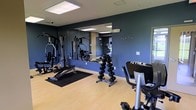 This fitness center is a thoughtfully designed workout space that provides everything needed for a comprehensive exercise routine. The room is finished with light hardwood-style flooring that contrasts attractively with deep navy-blue accent walls, creating a focused and energizing atmosphere. Large wall-mounted mirrors span a significant portion of the room, enhancing the sense of space and allowing users to monitor their form during exercise. The ceiling features recessed panel lighting that ensures the space is well-lit throughout. The facility is outfitted with a range of strength training equipment, including a multi-function cable and weight machine, an adjustable weight bench, and a full dumbbell rack with neatly organized free weights. Cardio equipment is also present, including a True-brand stationary bike, catering to both aerobic and anaerobic training needs. A dedicated mat area with a rubber floor mat provides a designated zone for floor-based exercises. The layout is practical and efficient, allowing multiple users to exercise simultaneously without feeling crowded. A coat hook rail on one wall adds a convenient amenity for gym-goers. Overall, the space is clean, modern, and functional, making it a well-rounded fitness facility.