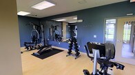 This fitness center is a thoughtfully designed workout space that provides everything needed for a comprehensive exercise routine. The room is finished with light hardwood-style flooring that contrasts attractively with deep navy-blue accent walls, creating a focused and energizing atmosphere. Large wall-mounted mirrors span a significant portion of the room, enhancing the sense of space and allowing users to monitor their form during exercise. The ceiling features recessed panel lighting that ensures the space is well-lit throughout. The facility is outfitted with a range of strength training equipment, including a multi-function cable and weight machine, an adjustable weight bench, and a full dumbbell rack with neatly organized free weights. Cardio equipment is also present, including a True-brand stationary bike, catering to both aerobic and anaerobic training needs. A dedicated mat area with a rubber floor mat provides a designated zone for floor-based exercises. The layout is practical and efficient, allowing multiple users to exercise simultaneously without feeling crowded. A coat hook rail on one wall adds a convenient amenity for gym-goers. Overall, the space is clean, modern, and functional, making it a well-rounded fitness facility.