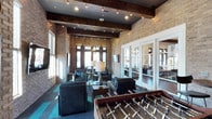 This inviting community lounge combines industrial-chic design with warm, welcoming character. Soaring ceilings are accented by rich, dark wood beams and strung with Edison-style pendant lights that cast a warm, ambient glow throughout the space. Exposed light brick walls lend a timeless, urban texture that serves as a striking backdrop for the room's modern furnishings.

The main seating area features plush, slate-blue upholstered chairs and sofas arranged around low coffee tables, creating comfortable conversation zones atop a teal-patterned area rug. A foosball table adds a recreational focal point, making the lounge ideal for social gatherings and casual entertainment. A dedicated dining or game table area near the entrance provides additional flexible space for group activities.

Large windows and glass-paned French doors allow natural light to fill the interior while offering views to adjacent spaces and the outdoors. A set of white-framed interior windows and doors connects this lounge to a neighboring room, enhancing the sense of openness and flow. The overall ambiance is sophisticated yet relaxed, balancing stylish design elements with functional, communal living in a space designed to bring people together.