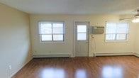 This single-floor space offers a clean and functional layout designed for comfortable everyday living. The main living area is highlighted by rich hardwood flooring that reflects natural light, contributing to a bright and welcoming atmosphere. Multiple windows fitted with horizontal blinds line the walls, allowing ample daylight to fill the room while providing privacy and light control as needed. A exterior-facing door with a glass panel insert provides direct access and adds an additional source of natural light. Climate control is well-addressed throughout the space, with a wall-mounted window air conditioning unit ensuring cooling comfort during warmer months, while electric baseboard heaters positioned along the walls provide efficient heating during cooler seasons. The walls are finished in a neutral, light tone that enhances the sense of openness and pairs well with the warm tones of the flooring. The overall ambiance is straightforward and practical, offering a move-in ready environment with essential features already in place. The space is well-suited for those seeking a no-frills, functional living arrangement with reliable climate control and a clean, easy-to-maintain interior.