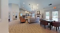 This single-floor community or professional space is thoughtfully designed to blend comfort with sophistication. The main lounge area serves as the centerpiece, featuring soaring vaulted ceilings with recessed lighting and an elegant chandelier that casts a warm glow throughout. A white brick fireplace with a rustic wood mantel anchors one wall, adding a welcoming focal point to the room. Neutral-toned carpeting complements the light gray walls, while an arrangement of plush sofas, accent armchairs, and a glass-topped side table create a refined, inviting atmosphere.

Natural light filters in through multiple windows and glass-paneled doors, keeping the space bright and airy. A bar-height seating area with stools is visible toward the rear, offering an additional gathering spot. The entryway transitions seamlessly from hardwood-style flooring to carpeted areas, with a framed artwork piece and a cushioned accent chair providing a polished first impression. Glass side panels at the entry allow views to the exterior, enhancing the sense of openness. Adjacent rooms, accessible through wide doorways, offer additional seating and private spaces, with hardwood floors and mirrored accents contributing to a consistently refined aesthetic throughout.
