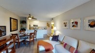 This single-floor residence offers a thoughtfully designed open-concept layout that seamlessly connects the living, dining, and kitchen areas into one cohesive and functional space. The living area is comfortably furnished with a light-toned sofa accented by colorful throw pillows, creating a warm and inviting atmosphere. A ceiling fan with a light fixture overhead provides both practicality and a subtle decorative touch. Light wood-style flooring runs throughout, lending a clean, unified look to the interior. The dining area sits adjacent to the living space and features a classic wooden dining set, ideal for casual meals or entertaining. The kitchen is finished with white cabinetry, a stainless steel refrigerator, and a range with an overhead microwave, all arranged around a convenient peninsula that doubles as a breakfast bar. Open shelving near the dining area adds a decorative element while providing additional storage. Neutral gray walls and a textured ceiling contribute to the understated, easy-to-personalize aesthetic. A hallway visible from the main living area suggests additional private rooms beyond, offering a clear separation between the shared living spaces and more secluded areas of the home. Overall, the space is bright, practical, and designed for comfortable everyday living.