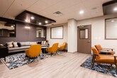 Located at 1 Woodbridge Center Dr in Woodbridge, NJ, this single-floor commercial lounge space combines contemporary design with functional comfort. The main lounge area is anchored by a large gray sectional sofa accented with colorful throw pillows, surrounded by bold mustard yellow accent chairs, orange and navy ottomans, and a leather armchair — all arranged atop a striking blue and gold patterned area rug. Light wood-look flooring runs throughout, providing a clean contrast to the darker design elements. A dramatic dark tray ceiling with recessed lighting defines the central lounge zone, while decorative wall sconces and framed mirrors add depth and elegance to the charcoal gray accent walls. A textured wood-panel feature wall serves as a focal point, with a wall-mounted flat-screen television integrated seamlessly into the design. The space also includes a well-appointed kitchenette featuring dark cabinetry, a white quartz countertop, stainless steel sink, built-in microwave, and an eye-catching mosaic tile backsplash in neutral tones. Structural columns are thoughtfully incorporated into the layout, and a glass-paneled door provides access to adjoining areas. The overall ambiance is modern, inviting, and professionally curated.