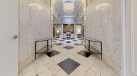 Located at 145 Granby Street in Norfolk, Virginia, this grand lobby makes a striking first impression with its refined classical architecture and upscale finishes. Floor-to-ceiling marble wall cladding in creamy white with subtle veining lines the corridor and main hall, creating a sense of timeless sophistication. The signature black-and-white diamond-patterned marble tile floor extends throughout, anchoring the space with bold visual contrast.

The lobby is organized into distinct zones that flow seamlessly into one another. The entrance vestibule is a stately, symmetrical passage flanked by wall sconces, tufted leather benches, and glass console tables, leading the eye toward the main hall. A skylight above the vestibule introduces natural light, brightening the polished surfaces below.

The main reception area is anchored by an elegant crystal chandelier suspended from a coffered ceiling with ornate crown molding. A rich wood reception desk with a glass base serves as the central workstation, accompanied by a pair of white upholstered accent chairs arranged for welcoming guests. Recessed lighting complements the chandeliers, ensuring the space is warmly and evenly illuminated throughout. Wall panel detailing and classical moldings reinforce the building's distinguished architectural character, making this lobby a sophisticated and functional gateway to the building.