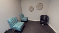 This entertainment room offers a welcoming and comfortable retreat designed with a coastal aesthetic in mind. The space is finished with neutral grey walls and dark speckled carpeting, creating a calm and grounded atmosphere throughout. Seating is a central feature, with a pair of teal-patterned armchairs and a sleek black leather swivel chair arranged to encourage conversation and relaxed viewing. A small round side table and a yellow upholstered ottoman add functional accent pieces to the layout. A wall-mounted flat-screen TV is positioned for easy viewing from all seating areas, accompanied by a light wood media console beneath it that holds decorative items, including a shorebird figurine and a seashell display, reinforcing the room's nautical theme. The walls are adorned with coastal-inspired artwork, including a framed triptych of shorebird watercolors and circular woven wall hangings that add texture and visual interest. A dark-framed door with glass panels provides access to the room, and a fire alarm and HVAC vent are neatly integrated into the space. The overall ambiance is relaxed and inviting, with thoughtful décor details that create a cohesive, beach-inspired environment suited for leisure and entertainment.