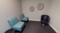 This entertainment room offers a welcoming and comfortable retreat designed with a coastal aesthetic in mind. The space is finished with neutral grey walls and dark speckled carpeting, creating a calm and grounded atmosphere throughout. Seating is a central feature, with a pair of teal-patterned armchairs and a sleek black leather swivel chair arranged to encourage conversation and relaxed viewing. A small round side table and a yellow upholstered ottoman add functional accent pieces to the layout. A wall-mounted flat-screen TV is positioned for easy viewing from all seating areas, accompanied by a light wood media console beneath it that holds decorative items, including a shorebird figurine and a seashell display, reinforcing the room's nautical theme. The walls are adorned with coastal-inspired artwork, including a framed triptych of shorebird watercolors and circular woven wall hangings that add texture and visual interest. A dark-framed door with glass panels provides access to the room, and a fire alarm and HVAC vent are neatly integrated into the space. The overall ambiance is relaxed and inviting, with thoughtful décor details that create a cohesive, beach-inspired environment suited for leisure and entertainment.