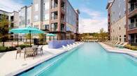 This amenity facility is part of a contemporary multi-story residential community and offers a well-rounded collection of health and recreational spaces. The outdoor area showcases a generously sized resort-style pool with crystal-clear turquoise water, lined with lounge chairs along a built-in sun shelf. A tiled water feature wall, black iron fencing, and teal market umbrellas with seating areas complete the polished outdoor environment, all set against a backdrop of open sky and surrounding greenery.

Inside, the fitness center is a standout feature, distinguished by soaring double-height ceilings, exposed silver ductwork, and an abundance of natural light streaming through large windows that frame outdoor views. A signature scallop-patterned carpet flows throughout the main workout area, which is equipped with treadmills, rowing machines, and multi-functional strength training systems. A dedicated spin room offers cycling bikes with digital screens alongside ceiling fans for ventilation.

A separate glass-enclosed studio provides a versatile flex space with hardwood flooring, mirrored walls, a large projection screen, and recessed lighting—ideal for yoga, group fitness, or media-assisted workouts. The overall design balances contemporary aesthetics with functional, high-quality fitness infrastructure.