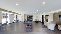 This single-floor amenity and recreation space offers a well-appointed environment designed for socializing and leisure. The open layout is anchored by a full-size billiards table with a blue felt surface and a sleek, contemporary pedestal base, positioned prominently at the center of the room. Dark hardwood-style flooring extends throughout, providing a rich contrast against the crisp white walls and smooth, recessed-lit ceiling above.

Seating options are thoughtfully distributed across the space, including a high-top bar table with chairs along one wall and a round dining table with chairs positioned toward the rear. A comfortable lounge chair is tucked into one corner alongside a billiards cue rack, catering to players and spectators alike. Large white panel doors and glass-panel black doors add architectural interest, while an open doorway leads into an adjoining room that appears to function as a bar or kitchenette area, complete with counter seating and pendant lighting.

Natural light filters in through glazed doors on one side of the room, enhancing the bright, airy ambiance created by the recessed lighting overhead. The overall design strikes a balance between modern minimalism and functional comfort, making this a versatile communal space well-suited for recreation and gatherings.