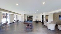 This single-floor amenity and recreation space offers a well-appointed environment designed for socializing and leisure. The open layout is anchored by a full-size billiards table with a blue felt surface and a sleek, contemporary pedestal base, positioned prominently at the center of the room. Dark hardwood-style flooring extends throughout, providing a rich contrast against the crisp white walls and smooth, recessed-lit ceiling above.

Seating options are thoughtfully distributed across the space, including a high-top bar table with chairs along one wall and a round dining table with chairs positioned toward the rear. A comfortable lounge chair is tucked into one corner alongside a billiards cue rack, catering to players and spectators alike. Large white panel doors and glass-panel black doors add architectural interest, while an open doorway leads into an adjoining room that appears to function as a bar or kitchenette area, complete with counter seating and pendant lighting.

Natural light filters in through glazed doors on one side of the room, enhancing the bright, airy ambiance created by the recessed lighting overhead. The overall design strikes a balance between modern minimalism and functional comfort, making this a versatile communal space well-suited for recreation and gatherings.